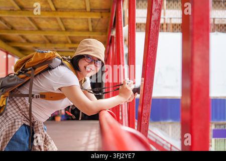 Smiling Asian Woman Tourist with Camera Leaning on Red Bridge Railing Stock Photo