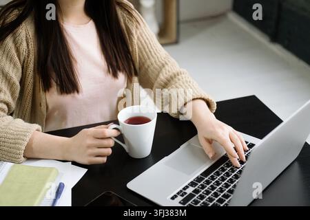 Close-up of woman's hands holding a cup of coffee while working on a laptop. Stock Photo