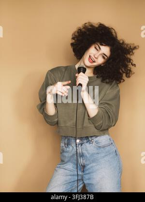 Cheerful curly-haired young woman sings songs into a microphone and dances. Stock Photo
