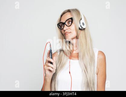 Senior woman wearing glasses listening to music with smartphone isolated on light grey background Stock Photo