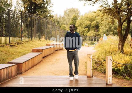 Portrait of a young african man dressed in a blue sweatshirt, holding a backpack, walking in the park on a summer day Stock Photo