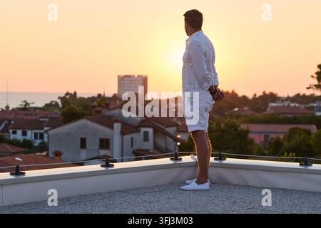 Man Standing on Rooftop Terrace Watching Sunset Over Seaside Town From Home Balcony Stock Photo