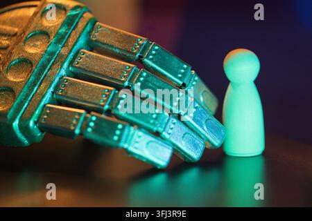 Metallic robotic hand extends towards a small wooden figure, highlighted by colorful blue and green lighting in a dark setting Stock Photo