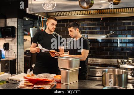 Two young adult chefs collaborating on menu planning and preparing food in a restaurant kitchen Stock Photo