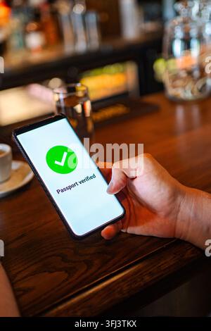 Hand holding smartphone displaying digital passport verified screen in a cafe Stock Photo