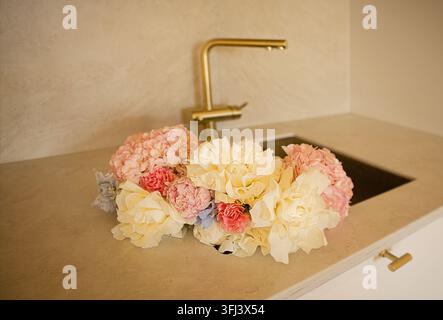 Pink and white artificial flowers are arranged on a sleek countertop beside a gold faucet in a contemporary kitchen environment Stock Photo