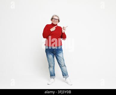 Smiling elderly gray-haired woman in red casual sweater pointing index finger aside up on mock up copy space Stock Photo