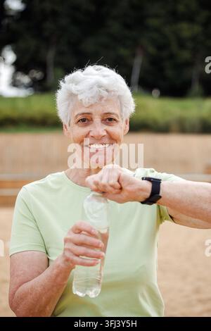 Senior woman with short grey hair drinking water after exercising, portrait in the park Stock Photo