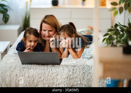 Mother and two daughters lying on bed watching content on laptop Stock Photo