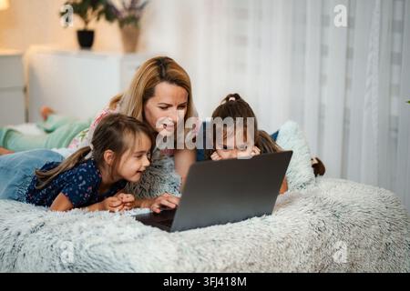 Mother and two daughters lying on bed using laptop for online learning Stock Photo