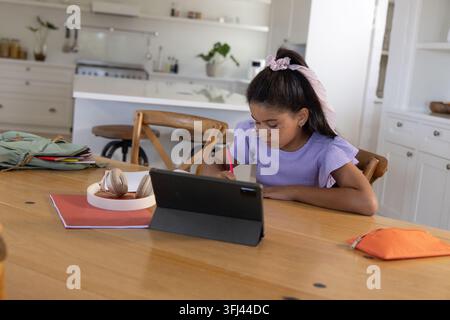 African American teen girl sitting at wooden table in kitchen writing with pink pencil using tablet. Student, remote learning, homework, notebook, hea Stock Photo