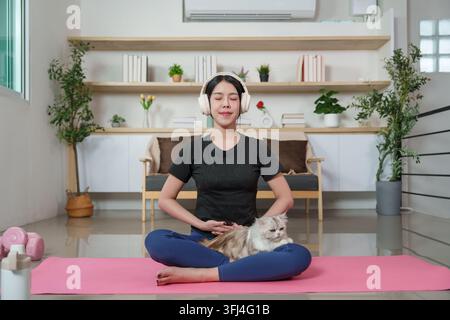 Asian woman meditating with wireless headphones and cat on yoga mat at home Stock Photo