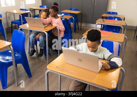 African American school-age children using silver laptops at classroom desks with blue chairs. Students, collaboration, technology, learning, educatio Stock Photo
