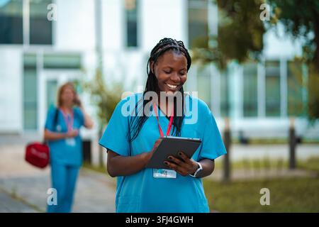 Smiling nurse in scrubs checking patient data on a tablet outside the hospital building Stock Photo