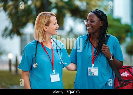 Diverse nurses smiling while walking together, discussing work outdoors Stock Photo