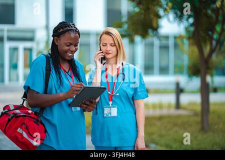 Two diverse medical professionals collaborating, using a digital tablet and smartphone outdoors Stock Photo