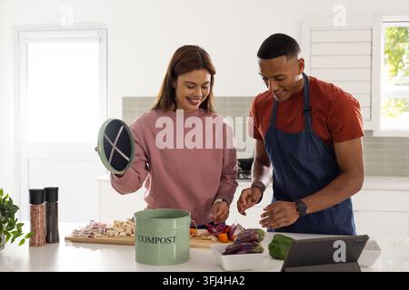 African couple chopping vegetables in home kitchen using compost bin with cutting board and tablet Stock Photo