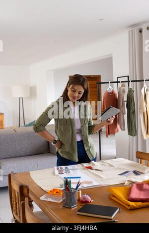 African woman in jeans standing in home studio reviewing sketches on tablet and arranging swatches Stock Photo