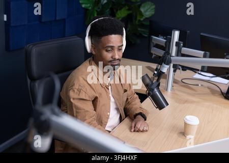 African American man sitting in studio at desk wearing headphones recording with mic, coffee cup Stock Photo