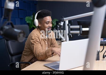 African American man speaking into mic, using laptop, wearing tan jacket and headphones in studio Stock Photo