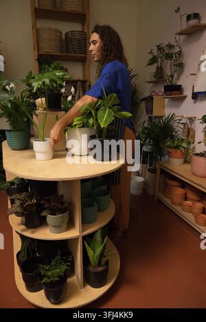 Nonbinary African American worker in blue top arranging plants on round display table in nursery. Greenery, terracotta, ceramic, Stock Photo