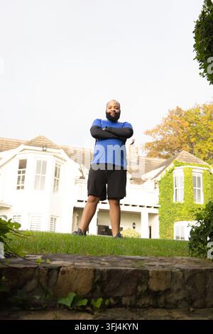 African American man standing on lawn by stone wall at two-story house wearing athletic shoes. Outdoor, frontyard, suburban, home, ivy, autumn, foliag Stock Photo