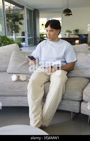 Adult man sitting on gray sectional sofa, holding smartphone and flipping notebook in living room. Modern, home, openplan, works Stock Photo