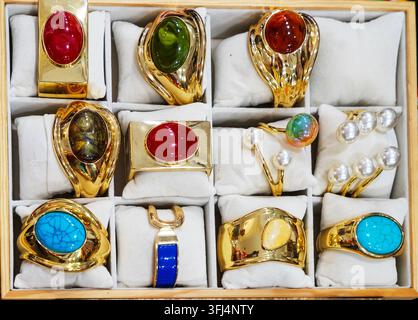 A flat-lay view of a wooden display case with white padded cushions showcasing a collection of luxury vintage-style statement jewelry. The assortment features wide gold-plated metal rings and cuff bracelets set with large polished oval gemstones in red, turquoise blue, emerald green, and amber brown, alongside elegant pearl-embellished multi-layered rings. Stock Photo