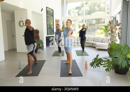 Diverse yoga class participants practicing standing pose on mats in bright living room with plants. Sunlit, wellness, group, instructor, interior, gar Stock Photo