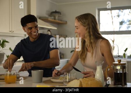 Diverse couple sitting at kitchen island preparing breakfast with French press, orange juice, bread. Home, morning, pair, conver Stock Photo