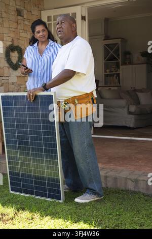 African American installer in toolbelt and homeowner holding phone checking solar panel on porch. Photovoltaic, technician, smartphone, entryway, ston Stock Photo