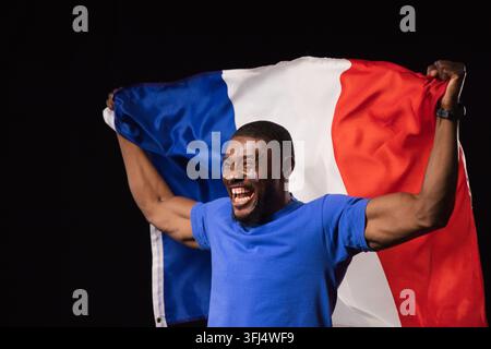 Midadult African man in studio holding tricolor flag, in blue tee, watch, face painted, cheering. Banner, celebratory, portrait, Stock Photo