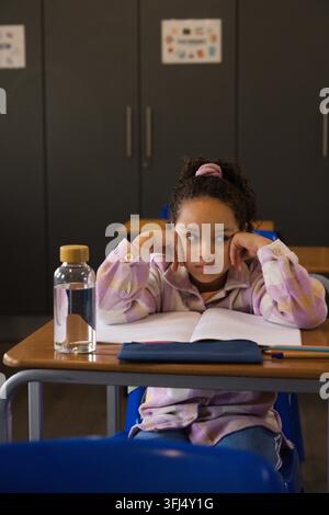 African American child in plaid shirt resting head on hands at classroom desk with notebook, bottle. Student, girl, focus, thoug Stock Photo