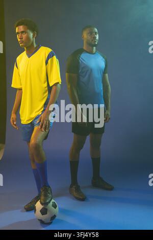 Two male athletes posing on blue seamless backdrop, wearing soccer jerseys and balancing ball. Duo, training, studio, lighting, haze, colored, sphere Stock Photo