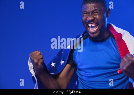 African American mid-adult man cheering, clenching fists, wearing American flag and shirt in studio. Triumph, celebration, victo Stock Photo