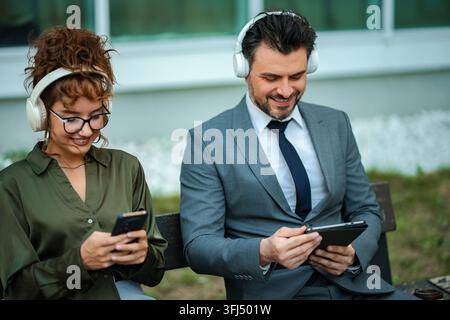 Two business colleagues wearing headphones, smiling and using digital devices outdoors Stock Photo