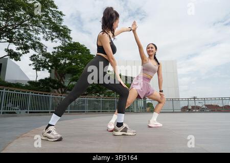Two active Asian female friends doing side lunge stretches and giving a high five outdoors on a city building terrace deck during morning exercise. Stock Photo
