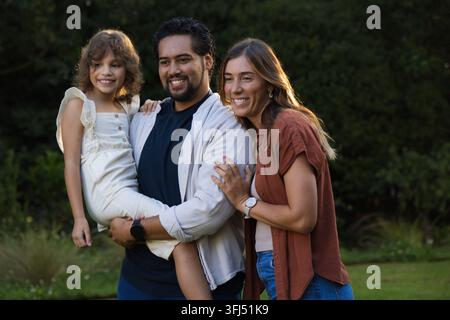 Diverse family smiling and posing in backyard, father holding child in white sleeveless dress. Trio, garden, lawn, shrubs, warmt Stock Photo