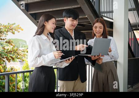 Group of three young Asian corporate professionals standing on a walkway, discussing work and pointing at a laptop screen together. Stock Photo
