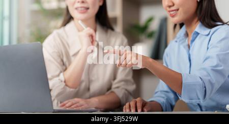 Two Asian female colleagues discussing online project details on a laptop screen while collaborating at a desk in a bright office. Stock Photo