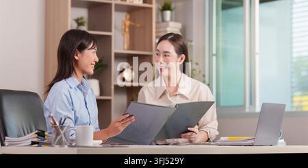 Two female colleagues discussing reports in document folders while sitting across a desk in a modern corporate office setting. Stock Photo