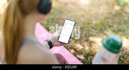 Over the shoulder view of a person holding a mobile phone displaying health activity data and heart rate statistics on a yoga mat in a park. Stock Photo