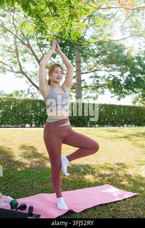 Asian woman practicing tree pose yoga balance outdoors on park lawn Stock Photo