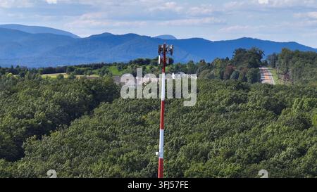 Tall red and white communication tower rises above dense green forest with residential buildings and mountains in background. Aerial view in Suburb of Stock Photo