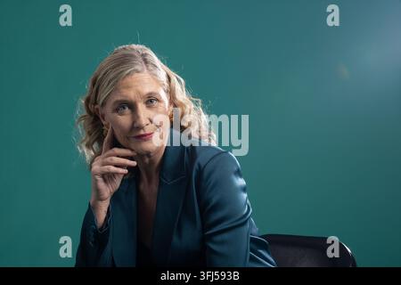Senior woman sitting, posing in studio on black leather chair, wearing teal satin blazer Stock Photo