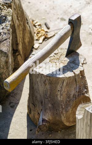 A weathered axe with a wooden handle rests on a tree stump used as a chopping block outdoors. Strong sunlight highlights the worn wood, metal head, ba Stock Photo