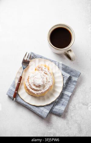 Freshly baked cinnamon bun drizzled white icing on a light background with a cup of black coffee, napkin and fork. A cozy Scandinavian breakfast conce Stock Photo