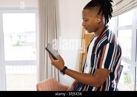 African American man standing by balcony wearing striped top and earbuds holding tablet, copy space Stock Photo