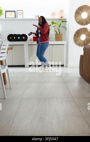 Adult woman standing in bright kitchen by counter holding speaker, wearing maroon jacket and jeans Stock Photo