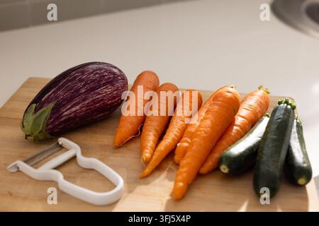 Wooden cutting board is holding striped eggplant, carrots, zucchinis, peeler on counter near sink Stock Photo